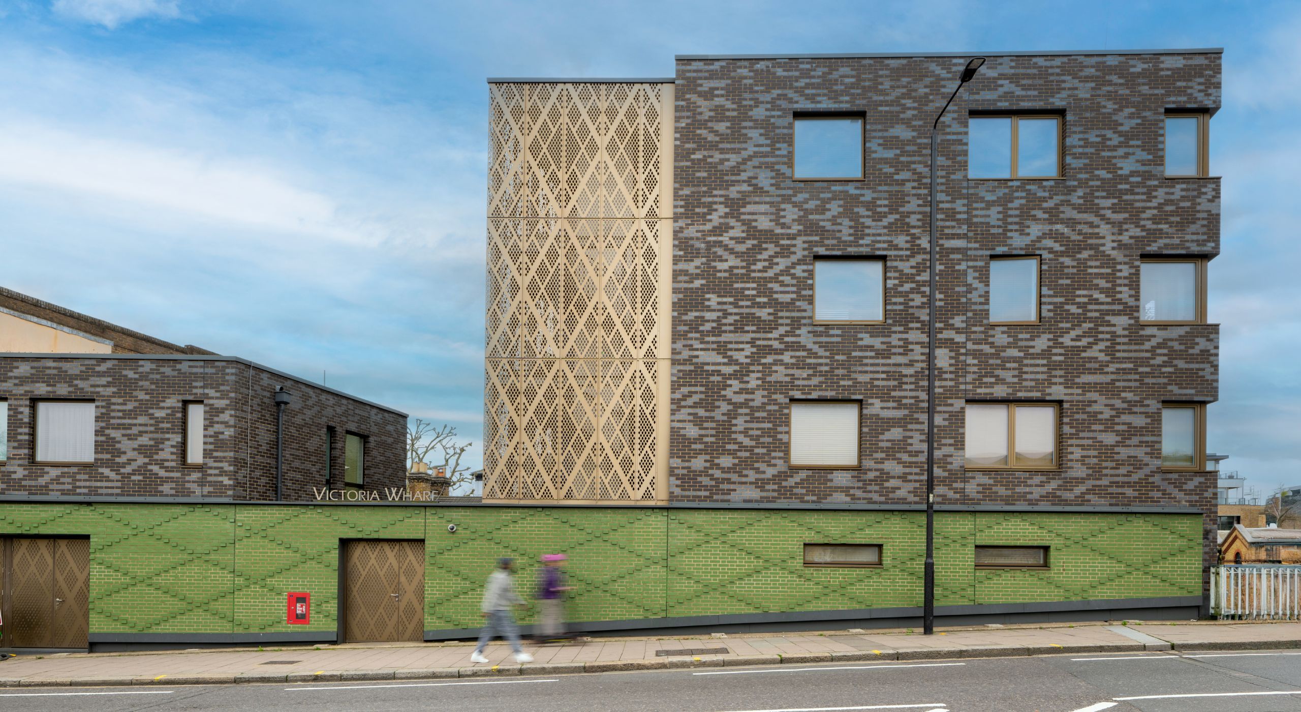A front image of a pattenred brickwork building that features windows edged with brown borders and a gold diamond perforated stair core.