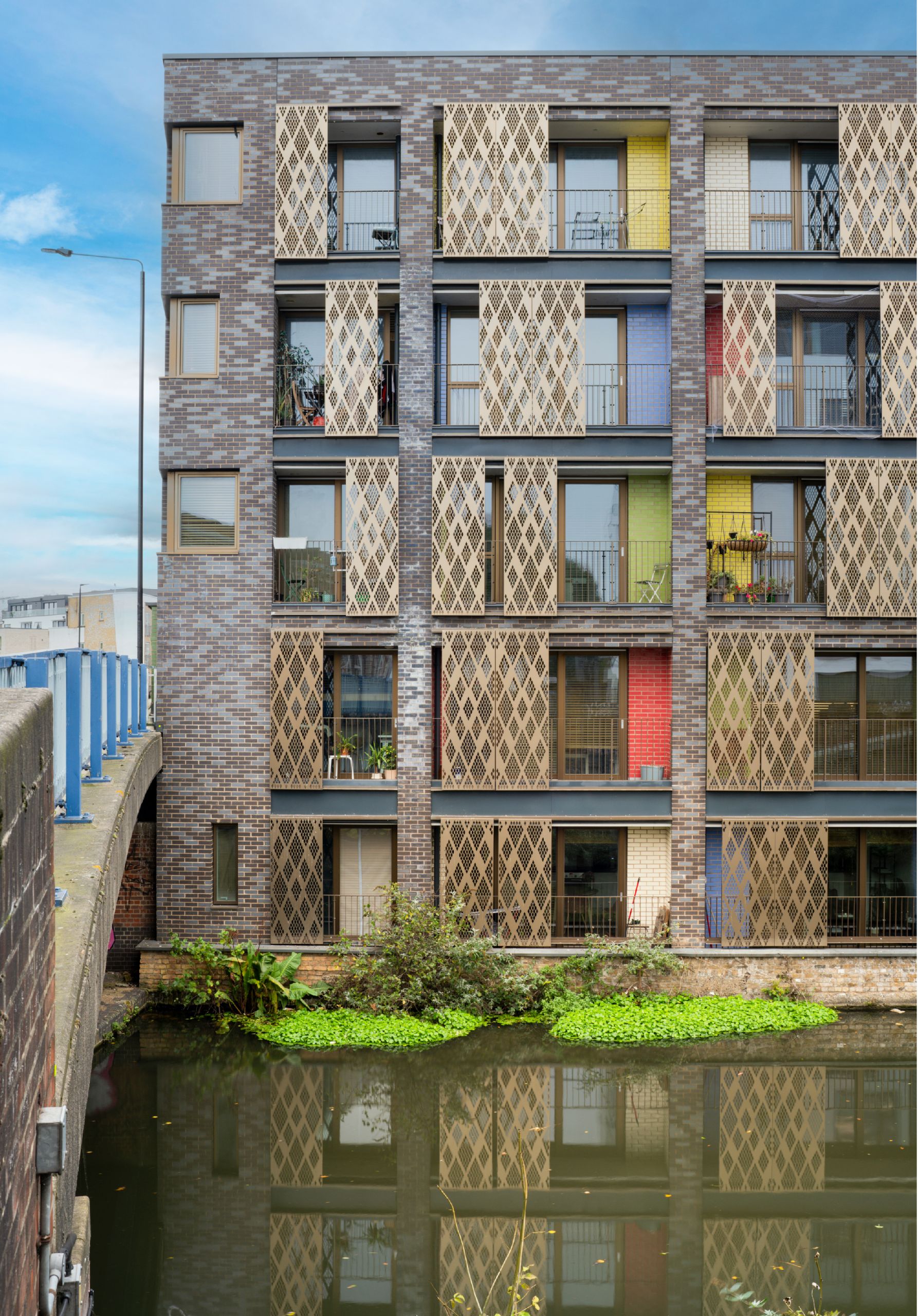 A patterned brickwork building with colourful bricked balconies and perforated gold panels on the balconies. The building is on a canal front.