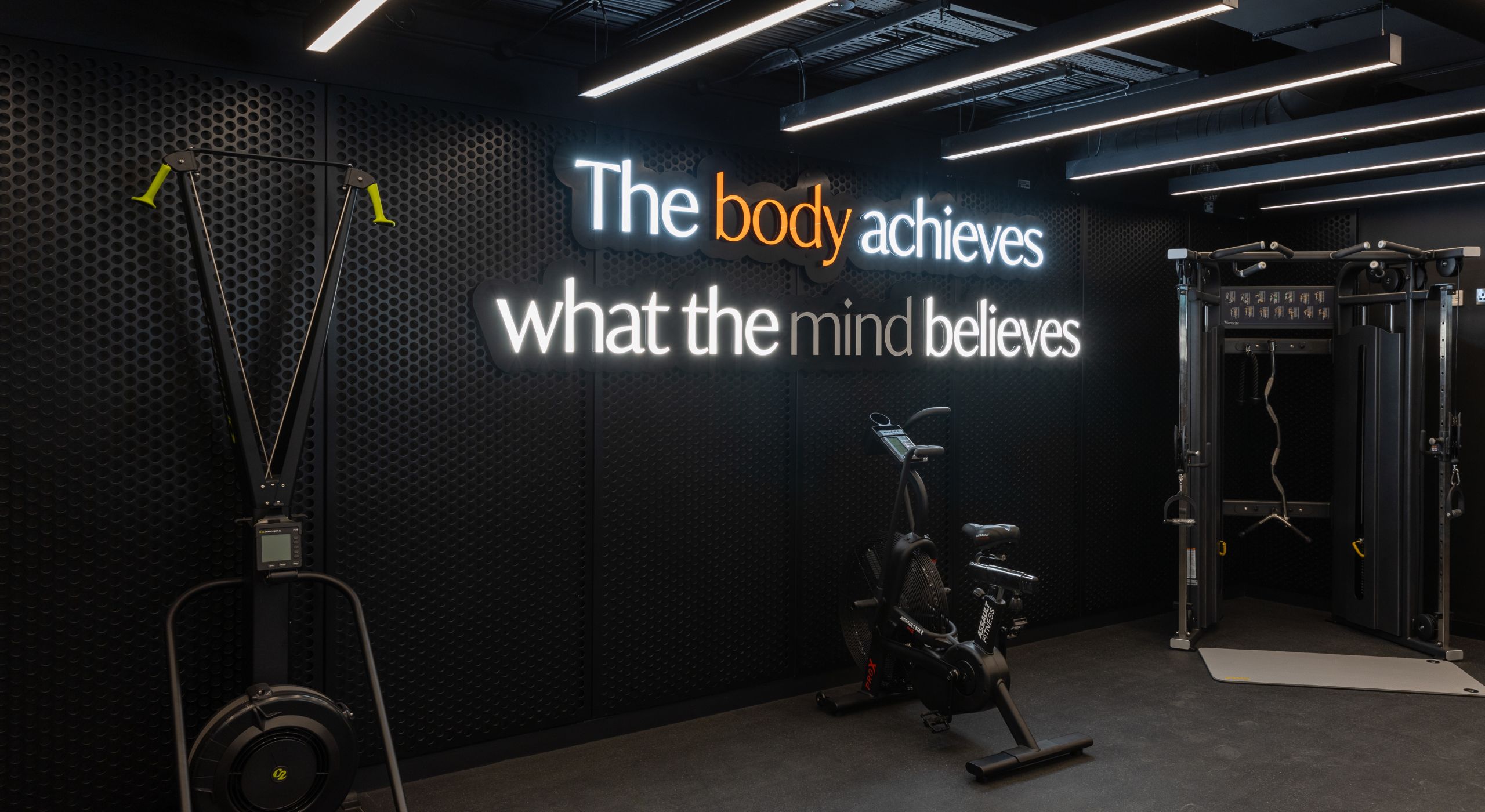 A gym with a black perforated metal wall with the words 'the body achieves what the mind believes'