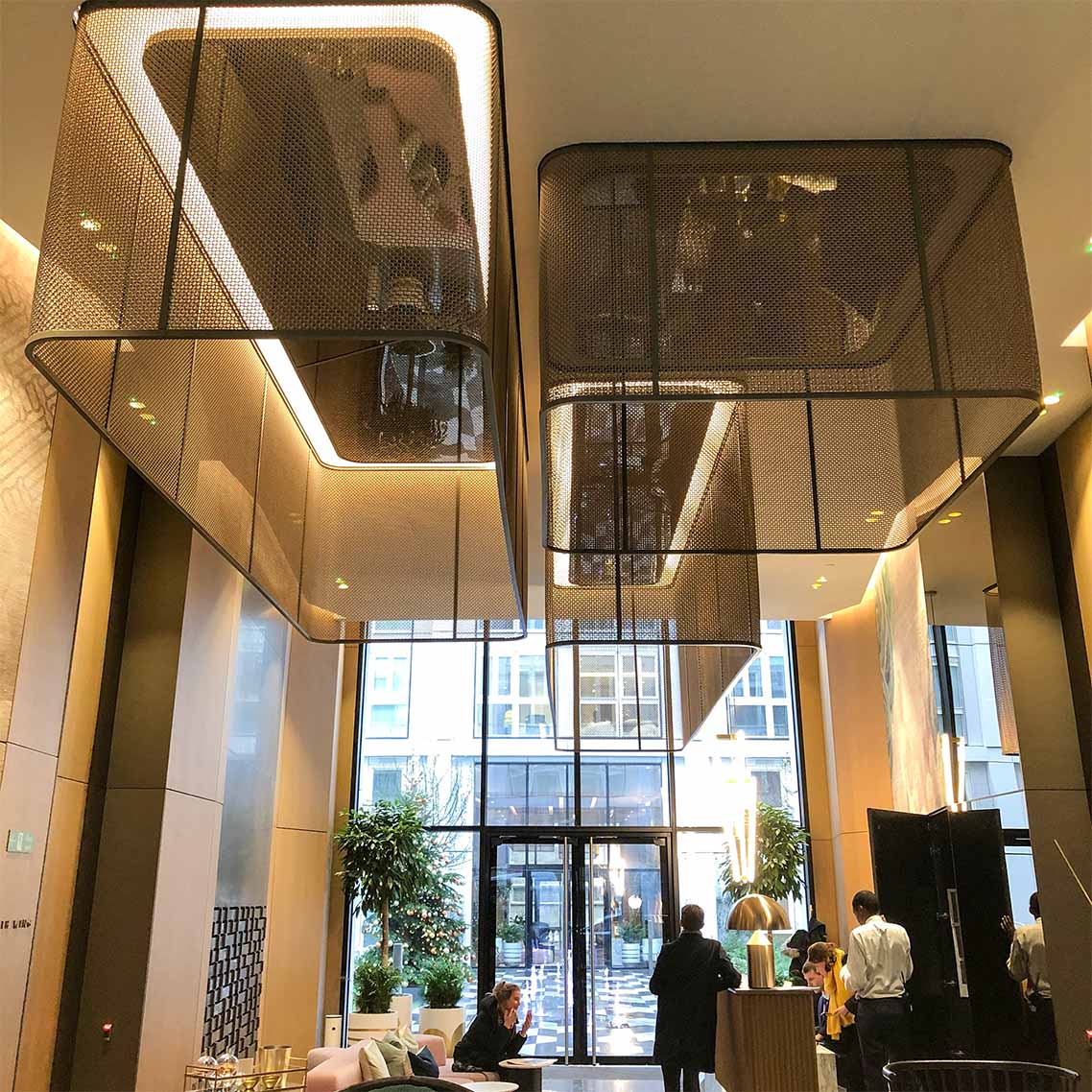 Reception area with large glass doors, with a golden gantry with a black trim in a gold woven mesh. with lights circling the top of the gantry on the ceiling.